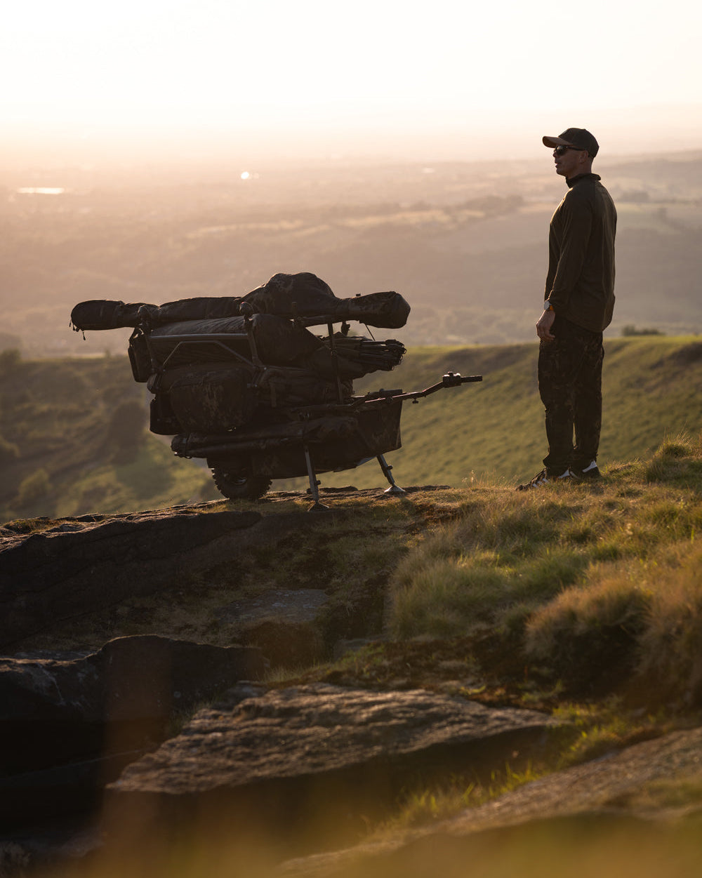 Person standing on a hill with a camera bag and tripod at sunset.